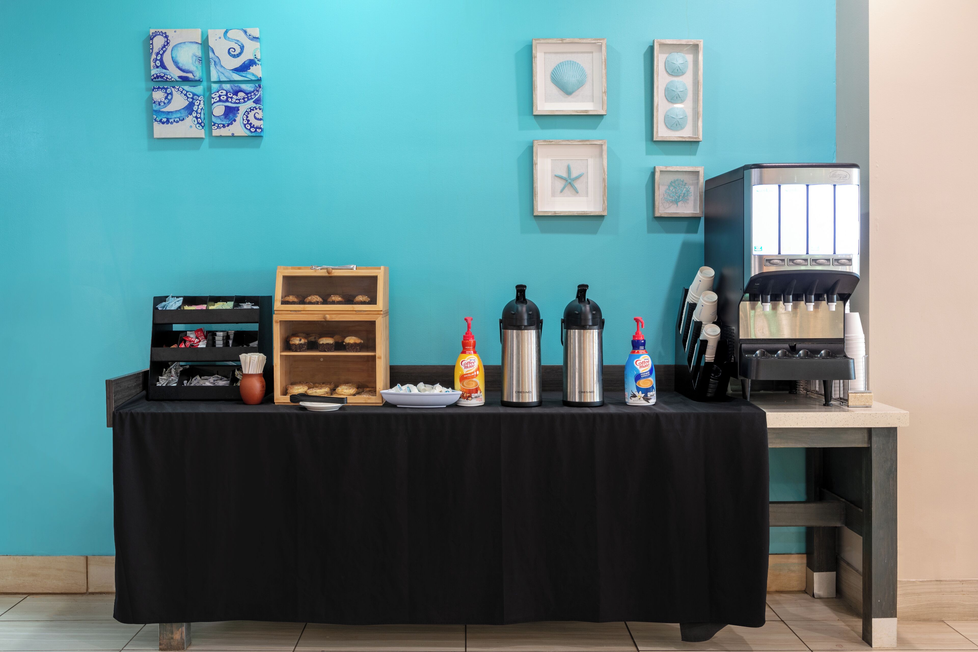 Uma mesa com comida, temperos e uma máquina de café em frente a uma parede azul-esverdeada.