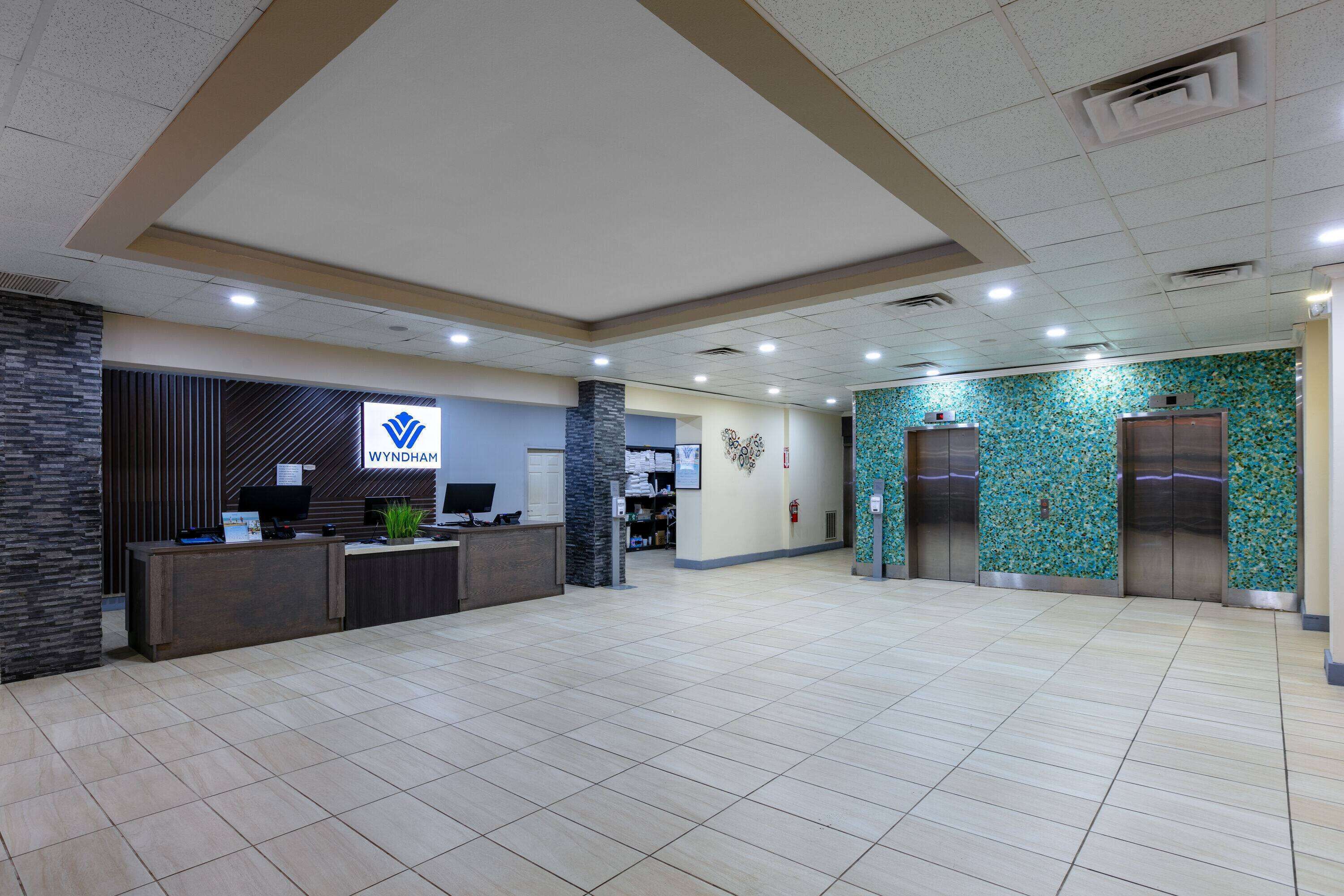 A reception area with a reception desk, a computer, and a logo for Wyndham. There are two elevators and a wall with a decorative pattern.