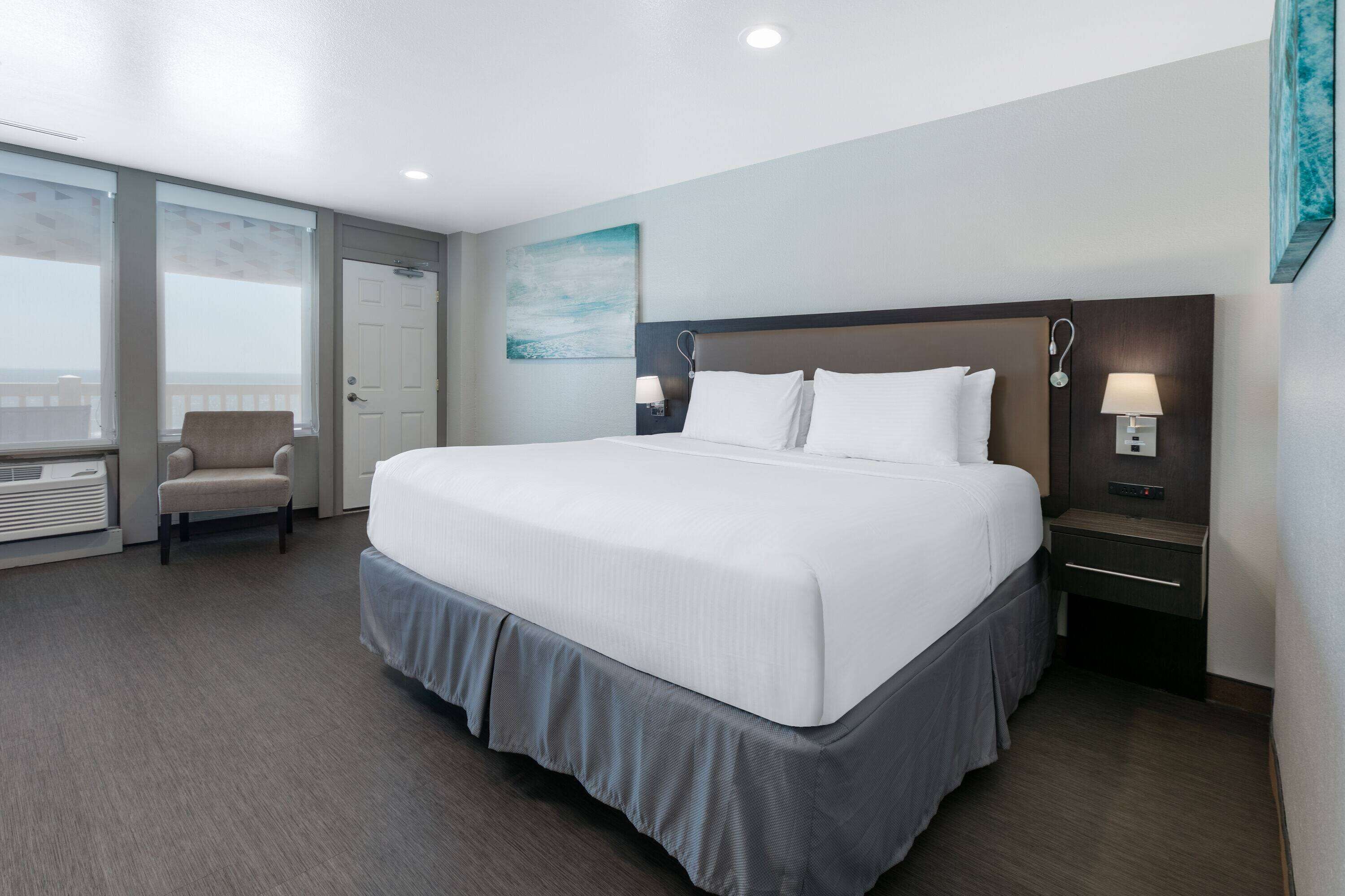 A hotel room with a large bed, a chair, a nightstand with a lamp, and a window with blinds.