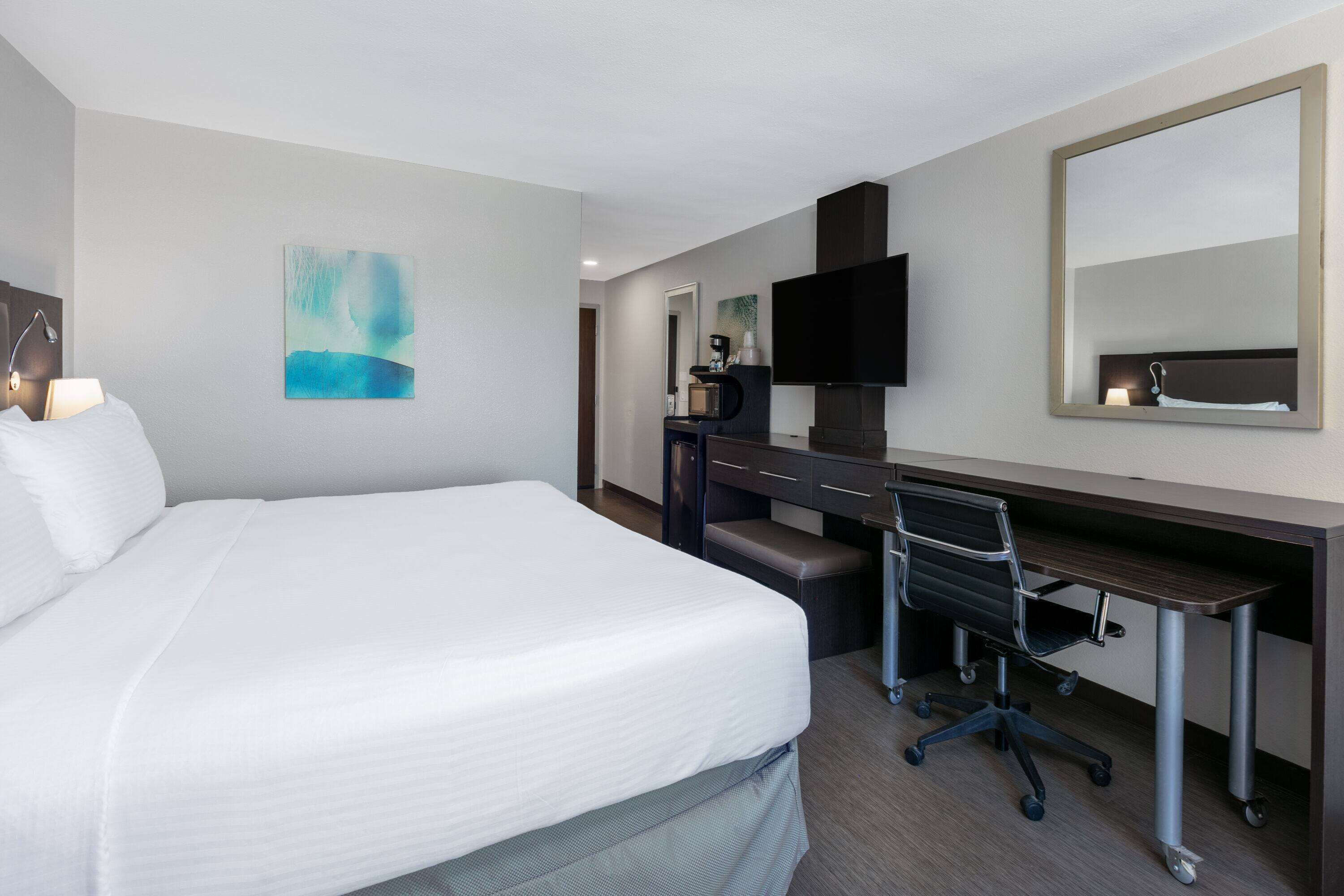 A hotel room with a large bed, a desk with a chair, a flat-screen TV, a coffee machine, and a mirror.