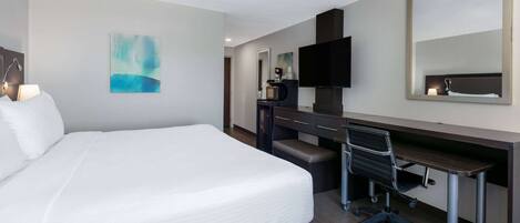 A hotel room with a large bed, a desk with a chair, a flat-screen TV, a coffee machine, and a mirror.
