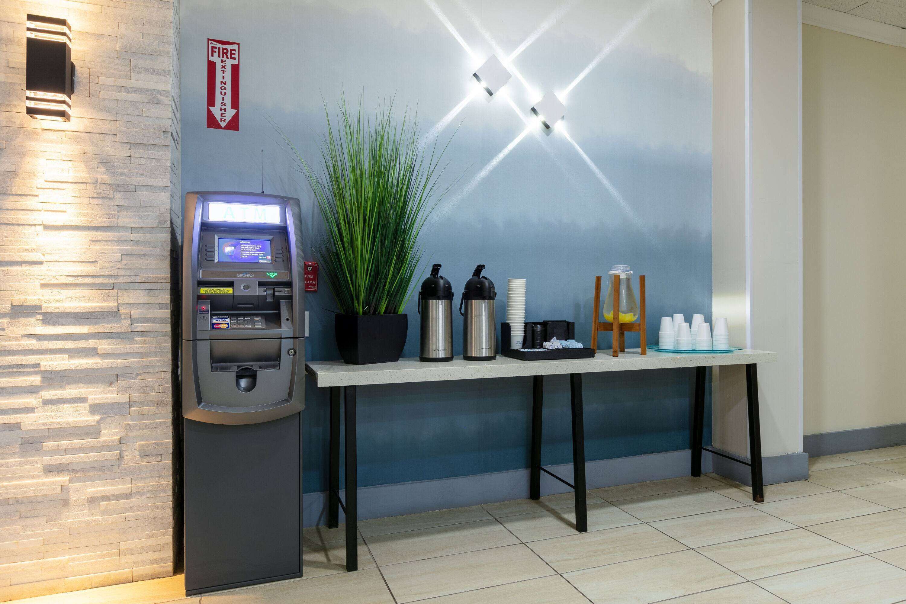 An ATM, a potted plant, and a table with cups and a coffee maker.