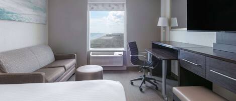 Ein Hotelzimmer mit Bett, Schreibtisch, Stuhl, Bank, Fernseher und einem Fenster mit Blick auf den Strand, sowie einer Klimaanlage.