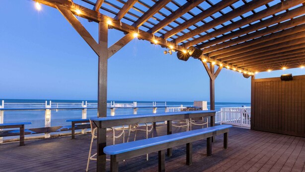 A wooden deck with benches and string lights, overlooking a body of water.