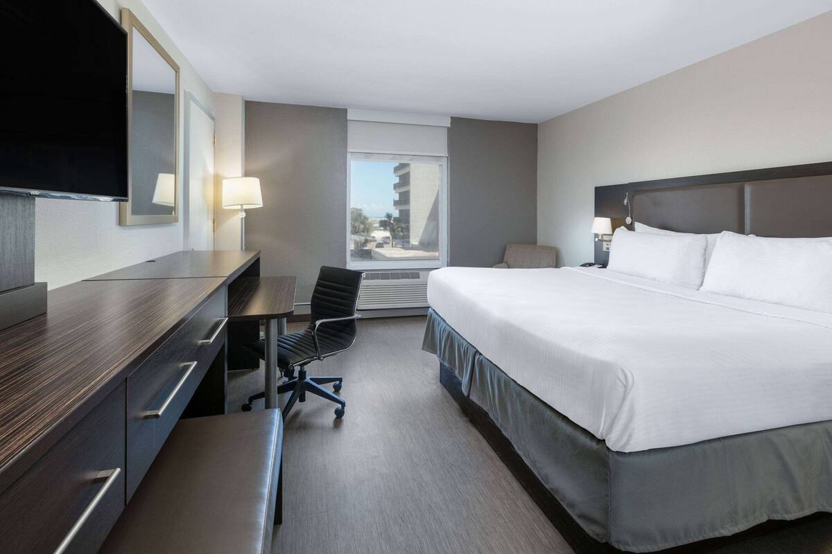 Een hotelkamer met een bed, bureau, stoel en een groot raam met uitzicht op de stad.