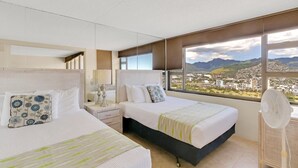 A hotel room with two beds, a large window with a city and mountain view, a bedside table with a lamp, and a standing fan. - Tower 2 Suite 3709 - QQP (Honolulu)
