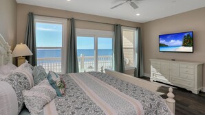 A bedroom with a large bed, a TV on the wall, a balcony with a view of the ocean, and a white dresser. - Pelican's Perch (Port St. Joe)