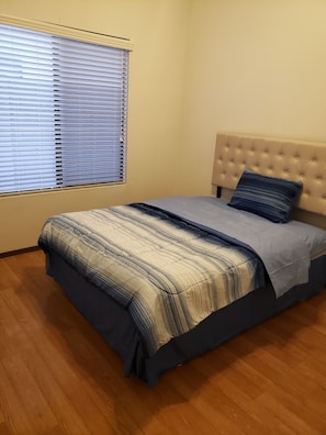 Ein Schlafzimmer mit Bett, Fenster mit Jalousien, einem Kopfteil und Holzboden.