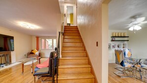 Uma escadaria leva a um segundo andar, uma área de refeições com mesa de vidro e uma cozinha com prateleiras.