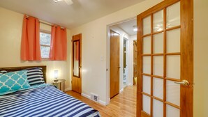 Quarto com cama, mesa de cabeceira, janela com cortinas, porta de madeira com painéis de vidro e um corredor que dá para outro quarto.