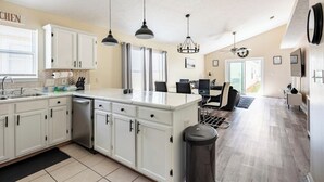 Une cuisine moderne avec des meubles blancs, un îlot central, des luminaires suspendus et un coin repas offrant une vue agréable.