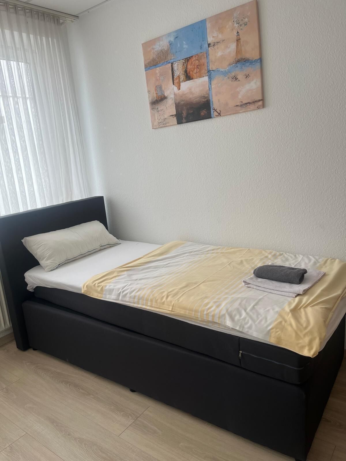 Ein ordentlich bezogenes Bett mit schwarzem Kopfteil, einer beige-weiß gestreiften Decke und einem gefalteten Handtuch.