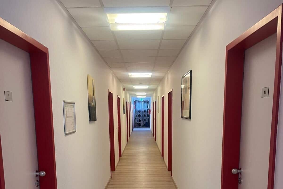 A long, narrow hallway with red-framed doors on both sides, wooden flooring, and overhead fluorescent lighting.