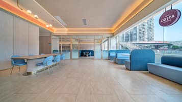 Um saguão de hotel moderno com uma mesa comprida e cadeiras azuis, uma área de estar com sofás azuis e uma grande janela com vista para a cidade.