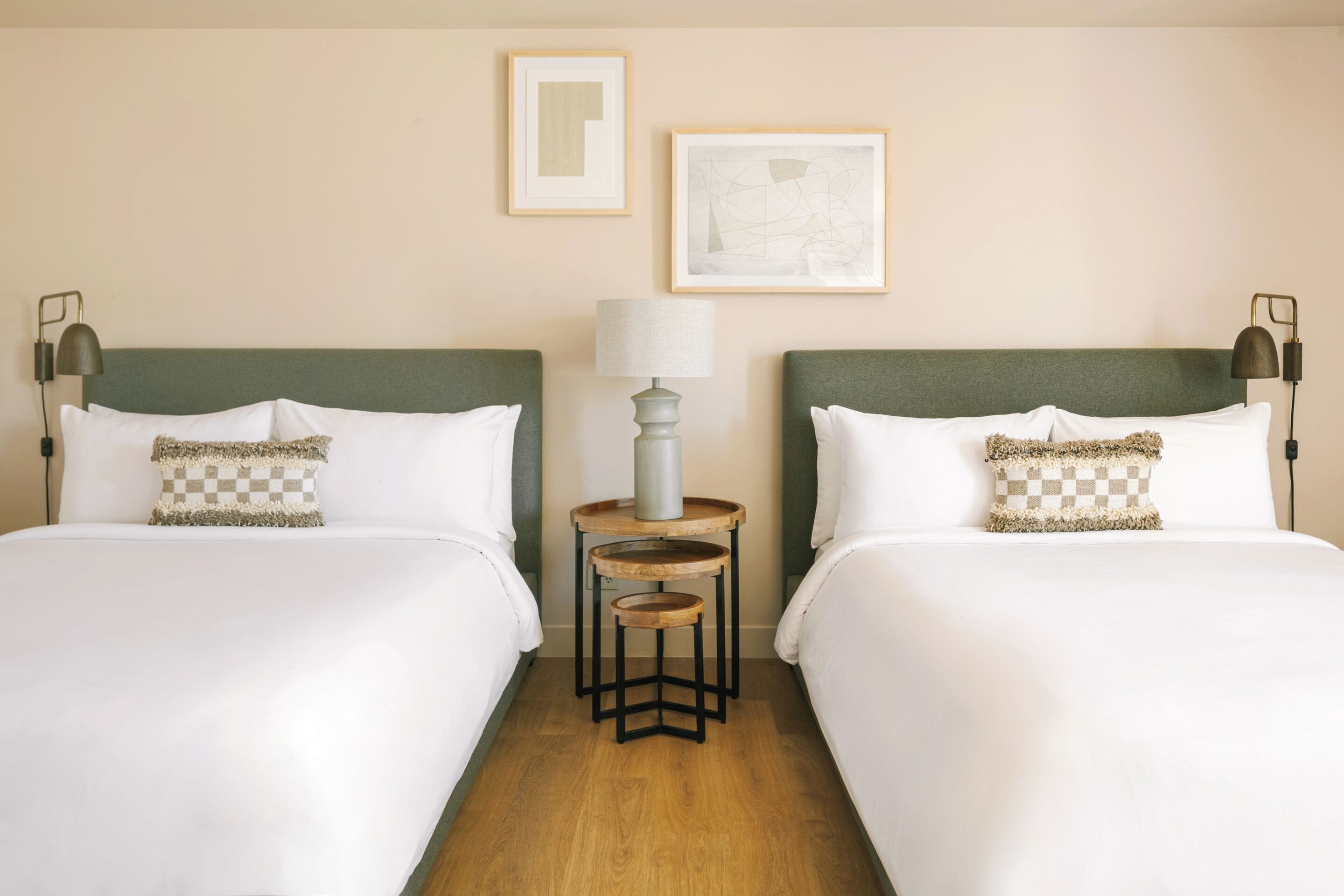 Ein Hotelzimmer mit zwei Betten, einem runden Tisch mit Lampe und zwei gerahmten Kunstwerken an der Wand.