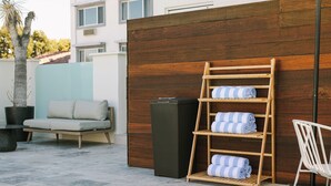 Un área de estar moderna al aire libre con un sofá, un estante de madera con toallas y un cubo de basura.