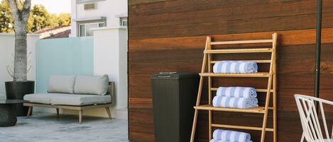 A modern outdoor seating area with a sofa, a wooden shelf with towels, and a trash bin.