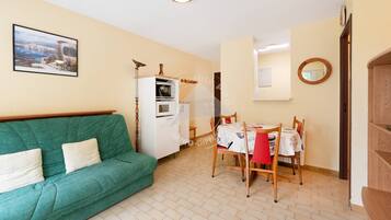 A cozy living room with a green sofa, a dining table with chairs, a white refrigerator, and a painting on the wall.