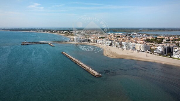 An aerial view of a coastal town with a beach, buildings, and a pier extending into the water. - AS21600-P2Cabin 200m from the beach and parking (LE GRAU DU ROI)