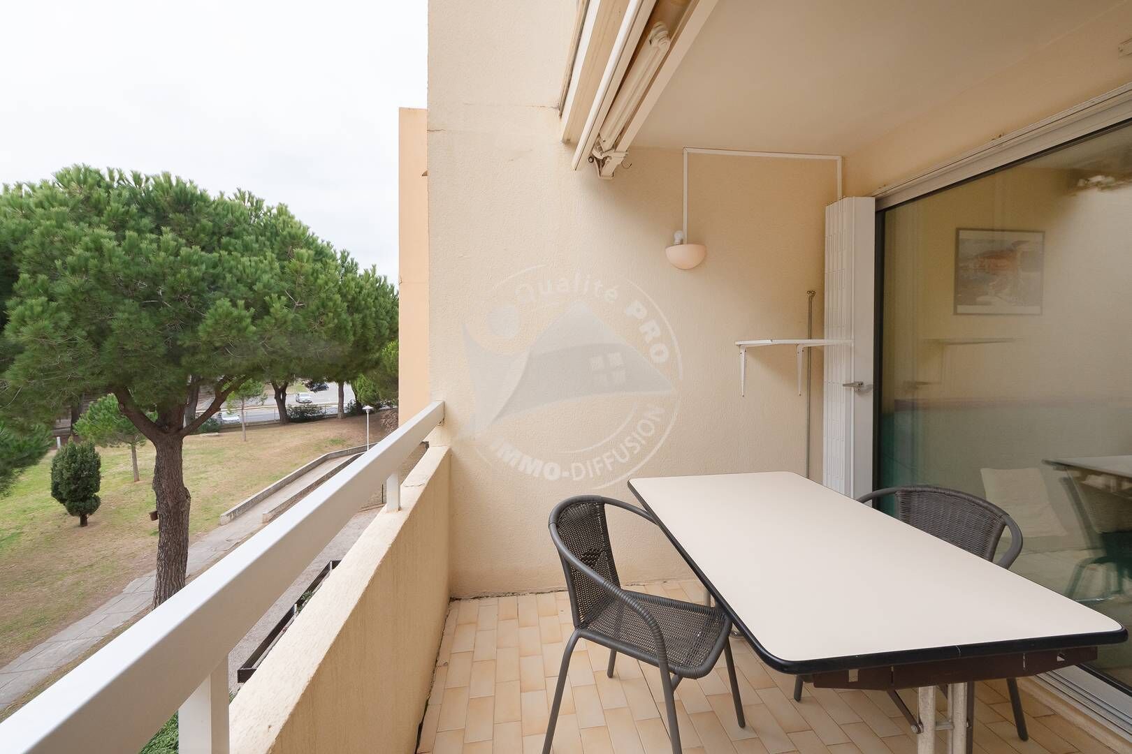 A balcony with a table and chairs, a glass door, and a view of trees and a car in the background.