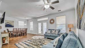 A spacious living room with a blue sofa, a dining area with chairs, a ceiling fan, and a large window with blinds. - ☛5 Minutes from the Beach★ Pool/ Hot Tub★ Stocked☚ (Destin)