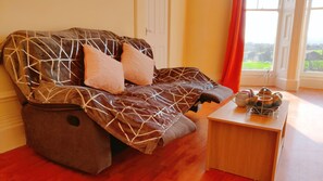 A massage chair with a geometric pattern, a small table with a teapot and cups, and a view of greenery through a window.