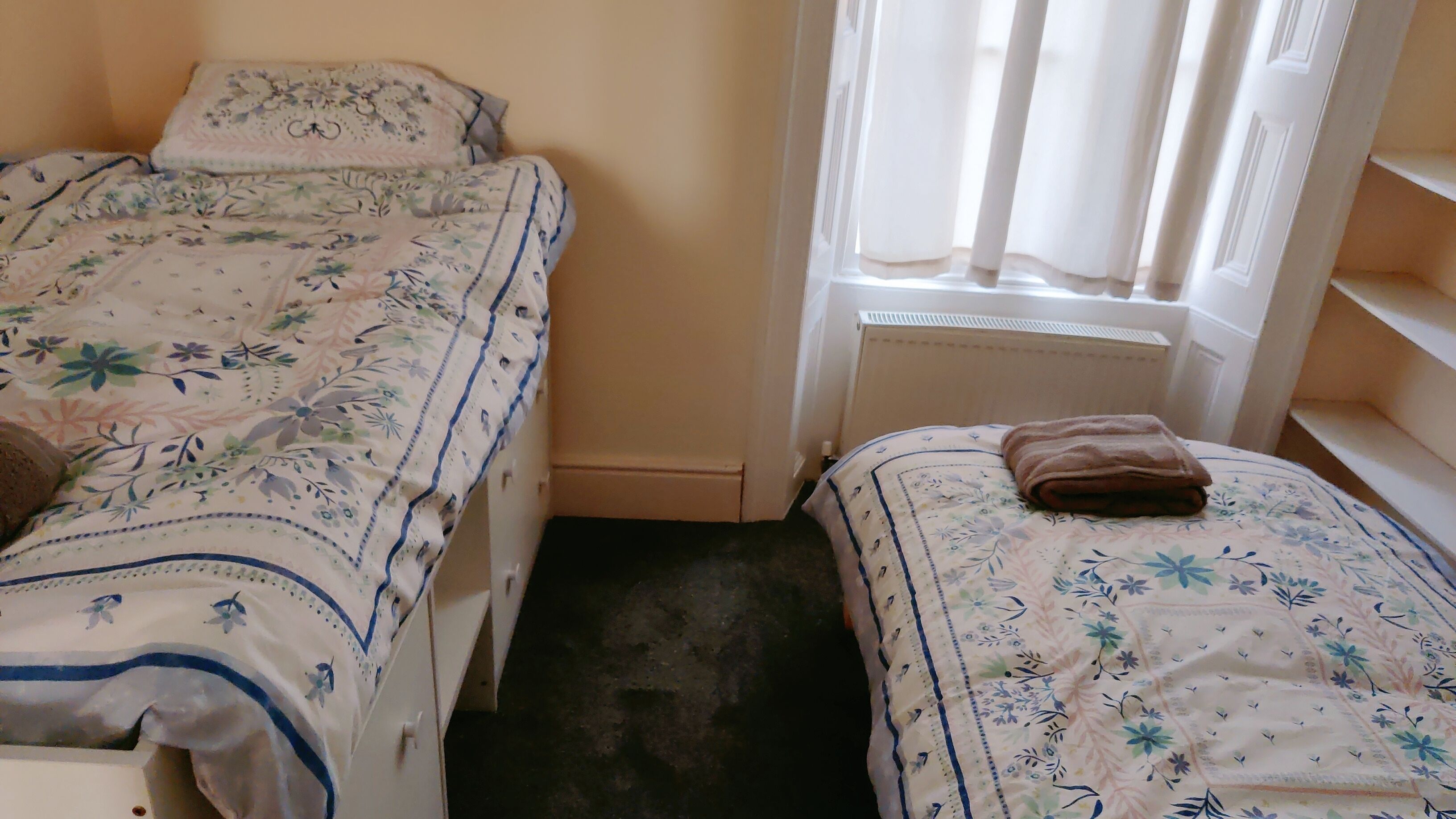 A small room with two single beds, each with floral-patterned bedding. There is a window with sheer curtains, and a brown towel is placed on one bed.