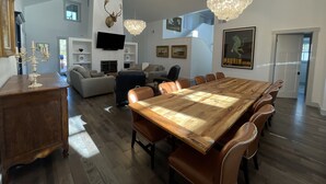 A spacious living area with a large wooden dining table, leather chairs, a chandelier, and a mounted deer head.