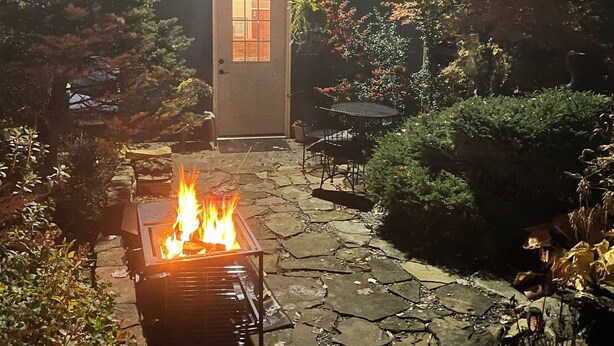A stone-paved patio with a fire pit, outdoor furniture, and a wooden building with a door and windows. - Cozy Buckhannon Cottage, Panoramic Views, Koi Pond & Close to Town (Buckhannon)
