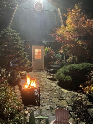 Eine terrasse mit Steinbelag, einem Feuerkorb, Gartenmöbeln und einem hölzernen Gebäude mit Tür und Fenstern.