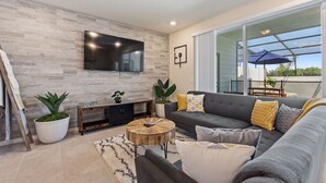 A modern living room with a flat-screen TV mounted on a textured wall, a grey sofa with multiple cushions, a round coffee table, and a potted plant.