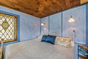 Une chambre avec un plafond en bois, un lit recouvert d’une literie à motifs, un vitrail et des lampes fixées au mur.
