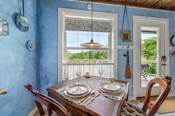 Un coin repas avec une table en bois dressée pour deux personnes, une fenêtre donnant sur un paysage verdoyant et un luminaire suspendu.