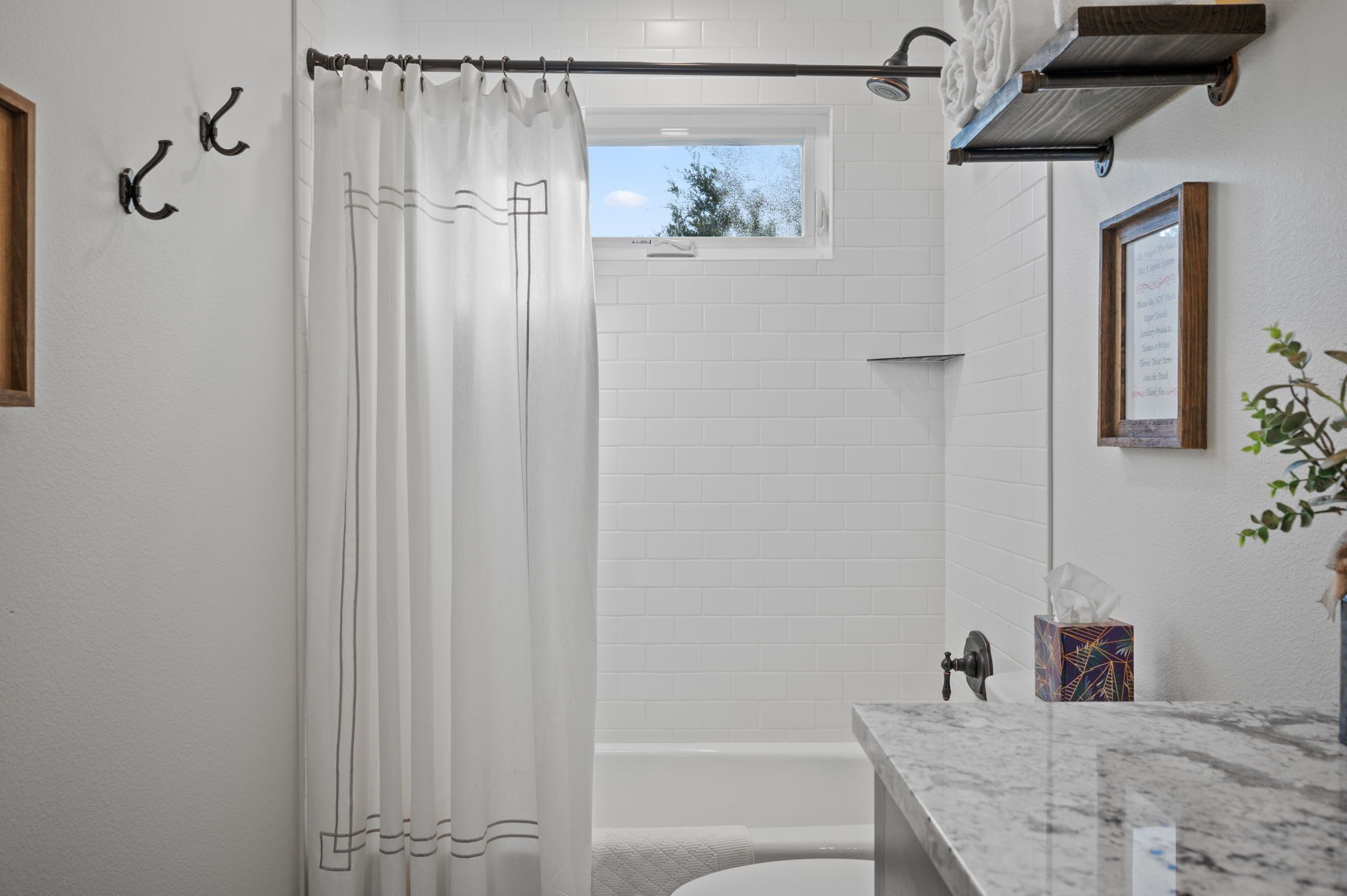 A bathroom with a white shower curtain, a window, a towel rack, a towel dispenser, a marble sink, and a framed picture on the wall.