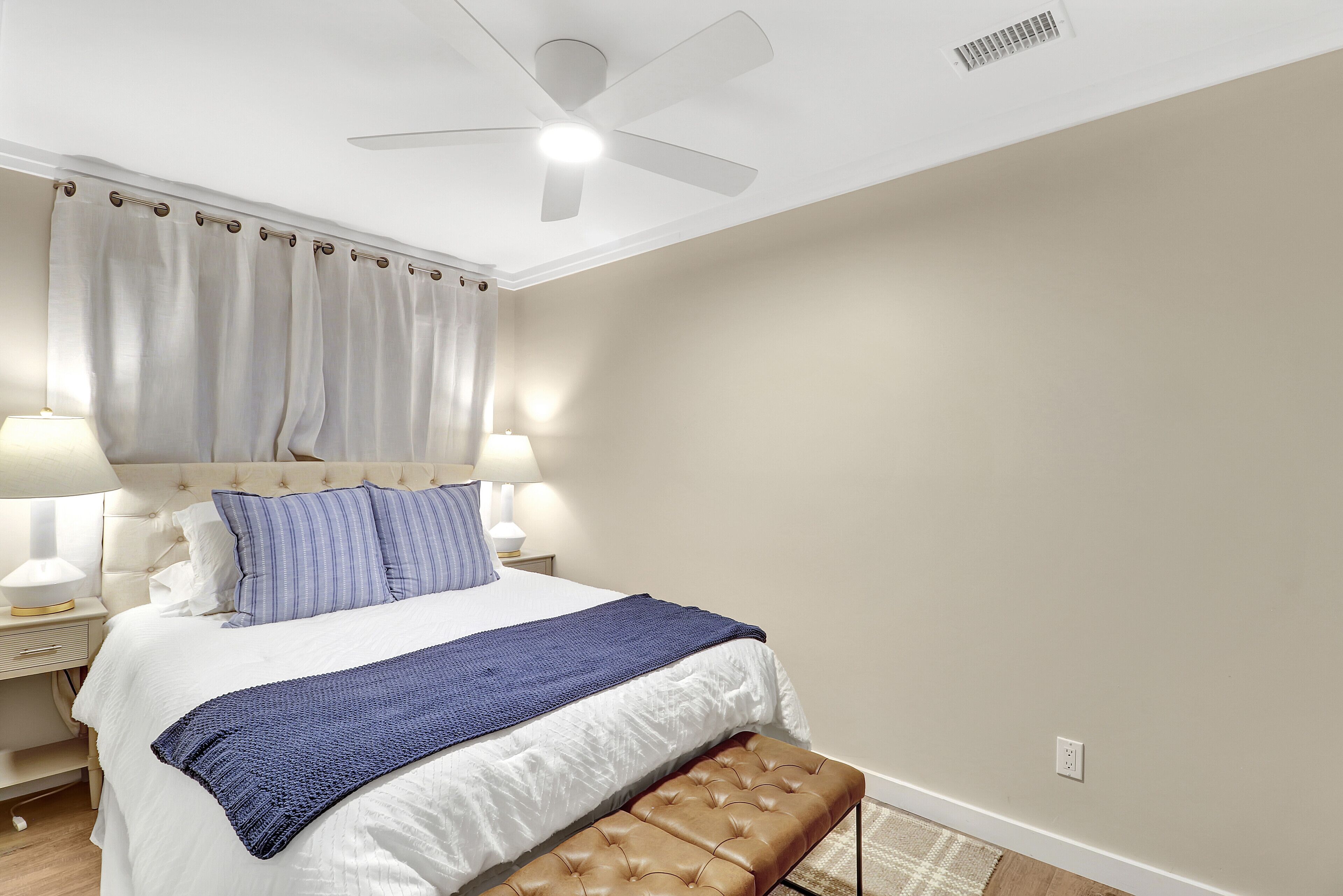 Un dormitorio con cama, lámparas de noche, ventilador de techo y ventana con cortinas.