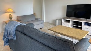 Ein modernes Wohnzimmer mit einem grauen Sofa, einem Holztisch, einem Fernseher auf einem Holzgestell und einer Lampe auf einem Beistelltisch.