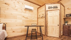 Une pièce de cabane en bois comprenant un lit, une petite table avec un tabouret, un appareil de climatisation, une porte avec un manteau accroché, et un coin cuisine avec des placards et un four à micro-ondes.