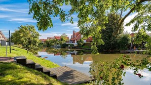 Ein ruhiges Gewässer mit einer hölzernen Pier, umgeben von Grünflächen und Häusern im Hintergrund.