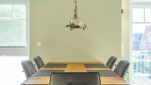 A dining room with a wooden table, six chairs, a hanging light fixture, and large windows.