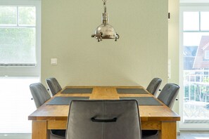 A dining room with a wooden table, six chairs, a hanging light fixture, and large windows.