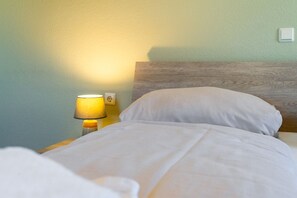 Uma cama com roupa de cama branca, cabeceira de madeira, mesa de cabeceira com abajur e tomada na parede.
