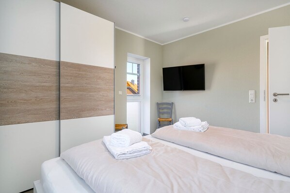 A hotel room with two beds, a flat-screen TV mounted on the wall, a wooden panel on the left wall, and a view of the outside through a window.