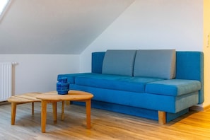 Um sofá azul, duas mesas de madeira e um vaso azul num quarto com teto inclinado e soalho de madeira.