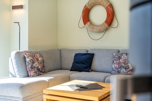 Un salon avec un canapé gris, des coussins aux motifs colorés, une table basse en bois avec une télécommande, et une bouée de sauvetage accrochée au mur.