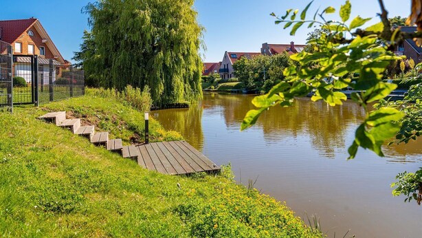 A riverside walkway with steps leading to a bridge, surrounded by greenery and residential buildings.