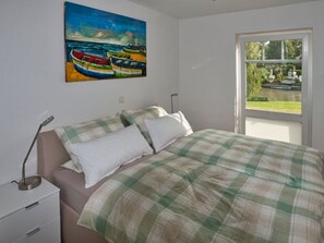 A bedroom with a bed, a nightstand, a painting of boats, and a window with a view.