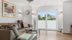 A spacious living room with a leather sofa, a coffee table, a painting on the wall, a clock, and a large sliding glass door leading to a balcony.