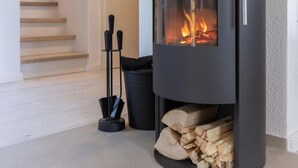 A modern wood-burning stove with a glass front, a chimney, and a stack of firewood beside it.