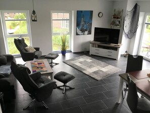 Ein modernes Wohnzimmer mit Fernseher, schwarzen Ledersesseln, einem Couchtisch und einem großen Fenster.