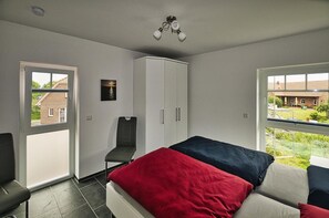 Ein Schlafzimmer mit Bett, Stuhl, Kleiderschrank und Fenster mit Blick auf ein Gebäude und Grünflächen.